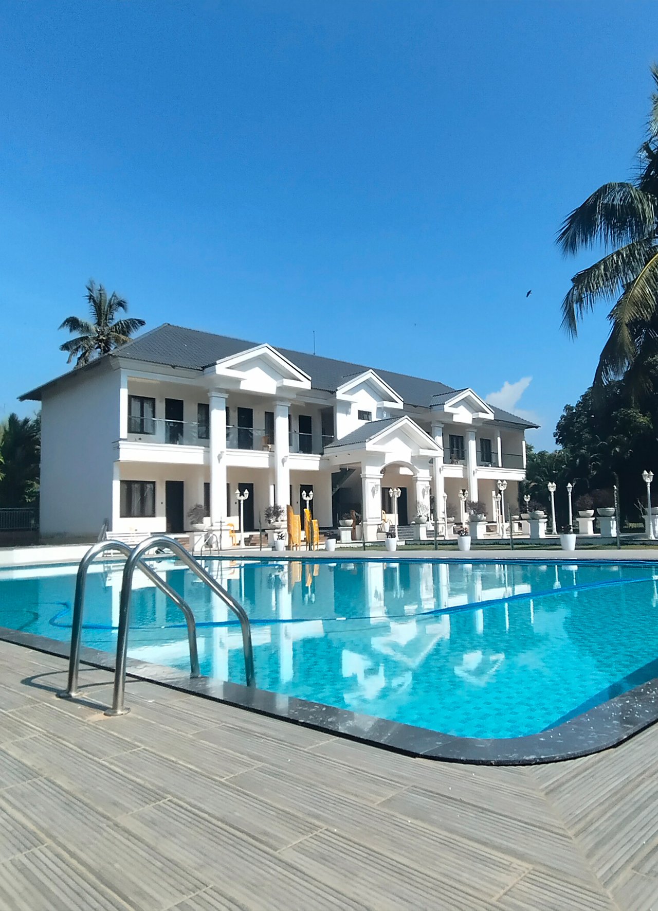 Wooden resort cottages with A-frame roofs and glass doors lining a curved, elevated walkway at sunset. Lush green lawn, tropical trees, and flowering bushes frame the scene.